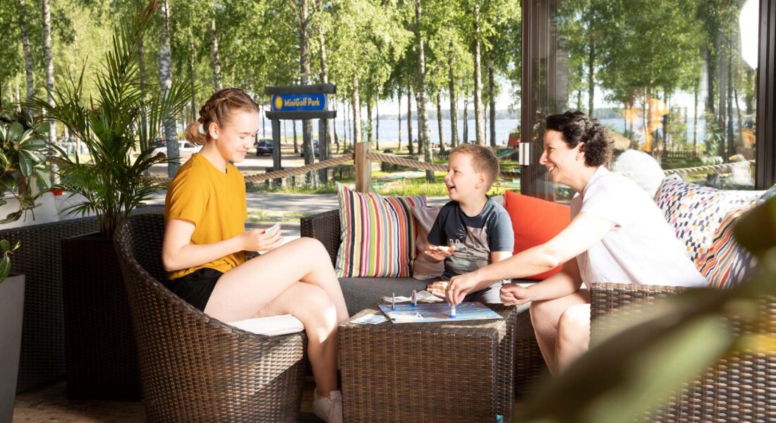 Bistro Santalahti terassi, kaksi lasta ja äiti pelaa lautapeliä, taustalla minigolf ja meri - Santalahti Resort - Kotka Finland